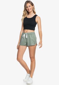 Top corto negro combinado con pantalones cortos verdes que presentan una cintura con cordón. La tela parece ligera y suave. Zapatillas blancas completan el look.