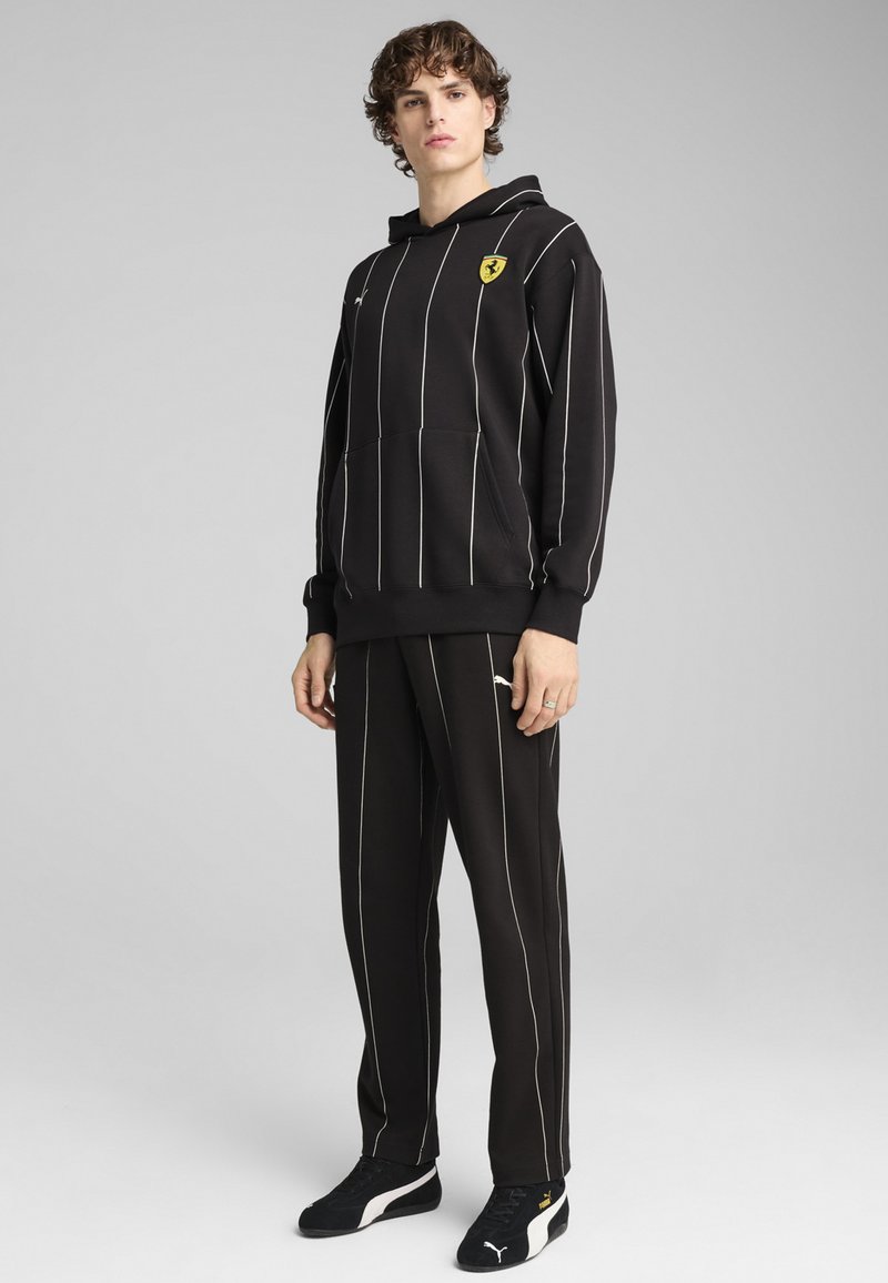 Crna dukserica s bijelim vertikalnim prugama, kenguru džepom i žutim logotipom. Uparen s odgovarajućim hlačama koje imaju slične pruge i naglašene logotipe.