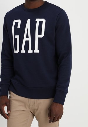 Mand iført navyblå GAP-sweatshirt og beige bukser, stående foran en ensartet lys baggrund.