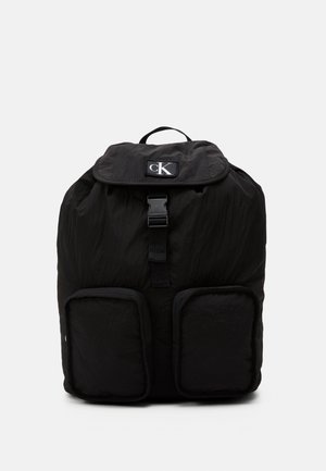 Czarny plecak Calvin Klein z klapką, zapięciem na klamrę, górnym uchwytem i dwoma przednimi kieszeniami na jednolitym jasnym tle.