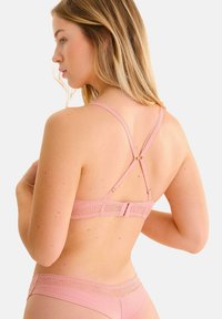 Soutien-gorge en dentelle rose clair avec des bretelles réglables et un fermoir à crochets. Présente un motif géométrique le long de la bande et une texture douce.