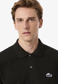 Polo nero con una trama tessuta, caratterizzato da una zip a due bottoni e un piccolo logo coccodrillo Lacoste colorato sul lato sinistro del petto.