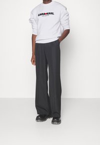 Sudadera blanca con el logo negro "CARL KARDASHIAN", combinada con pantalones de pierna ancha negros y zapatos gruesos negros. Tejido suave y corte relajado.