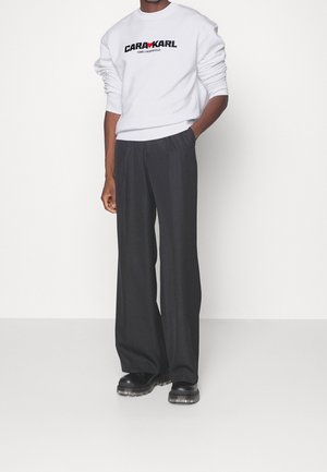 Sudadera blanca con el logo negro "CARL KARDASHIAN", combinada con pantalones de pierna ancha negros y zapatos gruesos negros. Tejido suave y corte relajado.