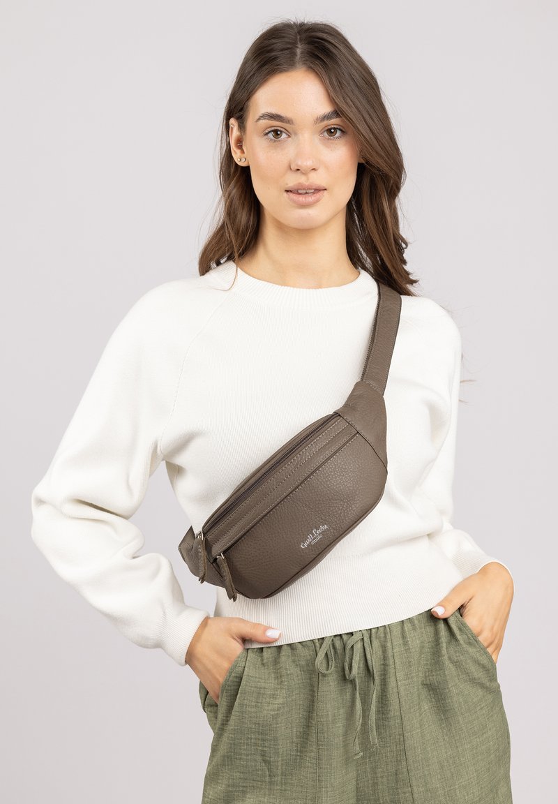 Braune Kunstleder-Umhängetasche mit strukturierter Oberfläche, einem Reißverschlussfach und verstellbarem Riemen, getragen über einem weißen Pullover und grünen Hosen.