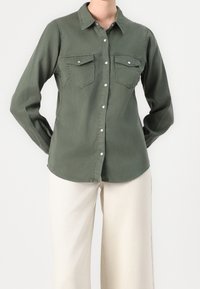 Chemise à manches longues à boutons en tissu vert olive, dotée de deux poches avant, d'un col classique et d'un ourlet droit. Associée à un pantalon large couleur crème.