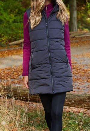 Femme portant un gilet sans manches matelassé noir sur un haut violet à manches longues, debout en extérieur sur un chemin recouvert de feuilles avec des arbres en arrière-plan.