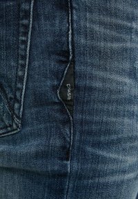 Jean en denim bleu foncé avec une texture subtilement délavée, cinq poches et une étiquette de marque visible cousue à l'intérieur de la poche.