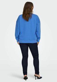 Sudadera azul de gran tamaño con puños y dobladillo acanalados, llevada sobre jeans oscuros ajustados y zapatos de tacón negros. Tela suave y cálida.