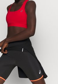 Top sportivo rosso con scollatura quadrata e logo grigio. Pantaloni corti neri con design aderente e accenti arancioni all'orlo, dotati di tasche laterali.