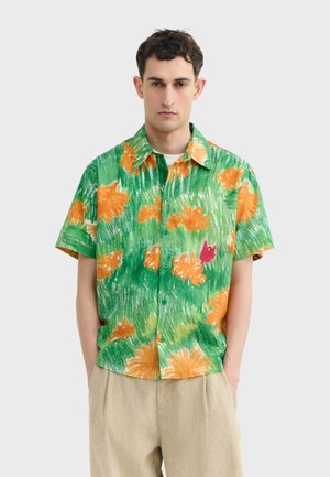 Mann trägt ein kurzärmliges grünes Hemd mit orange-rotem Blumenmuster, kombiniert mit beigen Hosen, vor einem schlichten Hintergrund stehend.