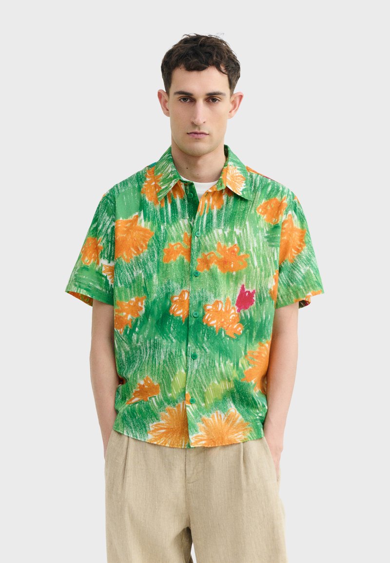 Homme portant une chemise à manches courtes verte à boutons avec un imprimé floral orange et rouge, associée à un pantalon beige, debout devant un fond uni.