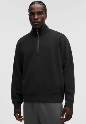 Mand med flettet hår har en sort sweatshirt med halvt lynlås og grå bukser på og står mod en ensfarvet lys baggrund.