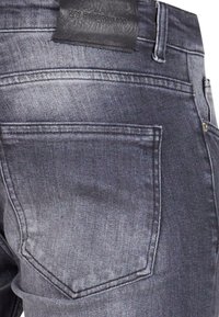 Gros plan sur la poche arrière d'un jean en denim gris foncé avec un patch en cuir noir portant l'inscription "2Y PREMIUM" au-dessus de la ceinture.