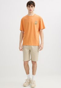 Joven de pie que lleva una camiseta naranja con un diseño floral, pantalones cortos beige, calcetines blancos y zapatillas beige sobre un fondo liso.