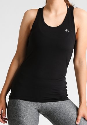 Kobieta ubrana w dopasowany czarny sportowy top na ramiączkach i szare teksturowane legginsy na jednolitym jasnym tle.