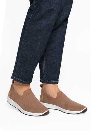 Feet wearing brown slip-on sneakers with white soles paired with dark blue cuffed denim jeans against a white background.