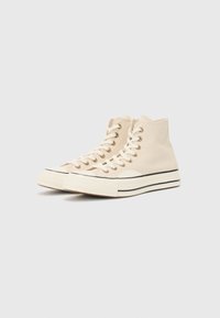 Beige high-top canvas sneakers with gold eyelets, white laces, black stripe on the sole, and a textured rubber toe cap.