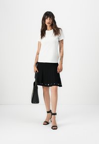 T-shirt blanc à manches courtes, jupe plissée noire avec des accents en œillets métalliques, sac à main noir et sandales à talons noires à lanières.
