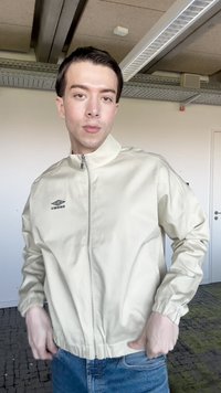 Jeune personne à l'intérieur portant une veste zippée Umbro crème et un jean bleu, debout avec les mains près des poches, expression neutre.