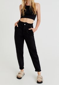 Svarta högmidjade byxor med en samlad elastisk midja, sidofickor, upprullade cuffs och en jämn textur, tillsammans med beige fluffiga sandaler.