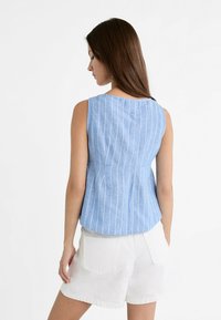 Žena s dlouhými hnědými vlasy, oblečená v modrém tílku bez rukávů s bílými vertikálními pruhy a bílých šortkách, stojící z profilu.