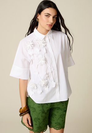 Mujer con cabello largo y oscuro lleva una blusa blanca de manga corta con aplicación floral y pantalones cortos verdes de ojal, accesorios con pulseras marrones gruesas.