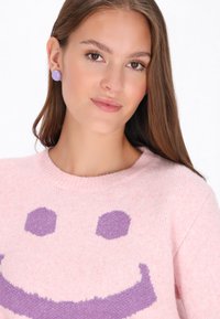 Rosa Strickpullover mit einem lila Smiley-Gesicht Design. Passende runde lavendelfarbene Ohrringe ergänzen den Look. Glatte Textur überall.