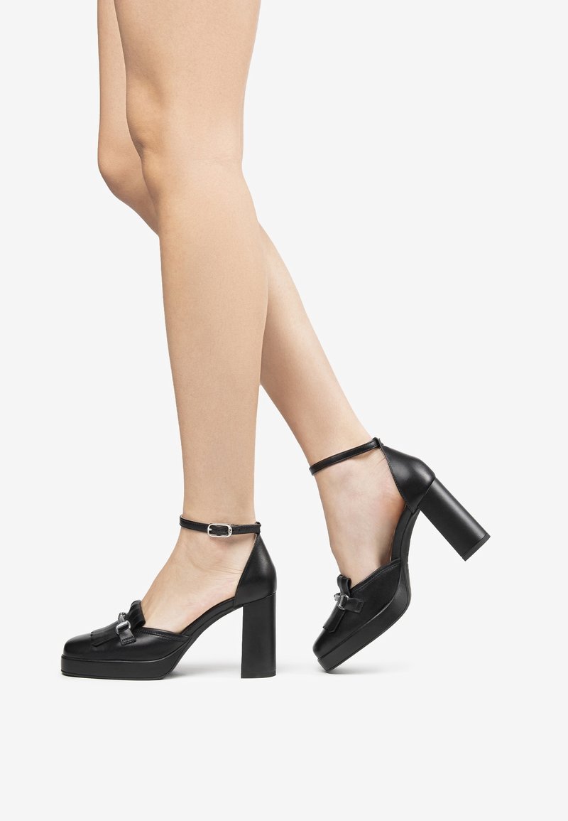 Jambes portant des talons compensés en cuir noir avec des brides à la cheville et des détails de boucle argentée sur fond blanc.