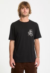 Man with light brown hair wearing a black t-shirt with a white cartoon character smoking a cigar on the left chest, paired with brown pants.