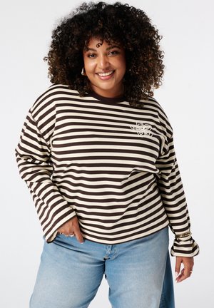 Femme souriante aux cheveux bouclés portant un t-shirt à manches longues rayé noir et blanc et un jean bleu clair, posant devant un fond blanc.
