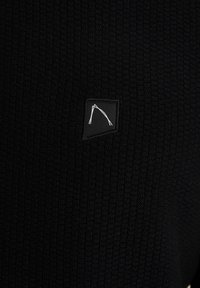 Tessuto nero texturizzato con un motivo a costine, dotato di un piccolo logo ricamato in bianco su uno sfondo quadrato nero.