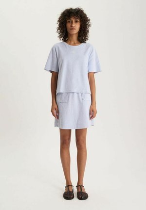 Femme aux cheveux bouclés portant un haut bleu clair à manches courtes et une jupe assortie avec des poches sur le devant, debout devant un fond uni.