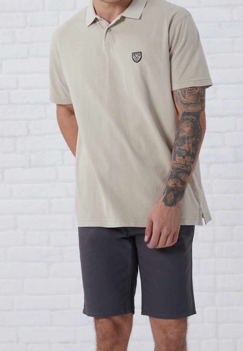 Homme portant un polo beige et un short gris foncé, affichant une manche de tatouage détaillée avec des motifs religieux et floraux sur le bras droit.