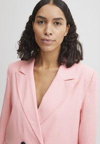 Rosa blazer med dyb V-hals, strukturerede skuldre, skarpe klapper og en glat tekstur. Har dobbeltknaplukning og en skræddersyet pasform.