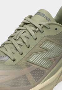 Κοντινή λήψη ελαφρώς πράσινου αθλητικού παπουτσιού με υφασμάτινη υφή, σταυρωτά κορδόνια και μεγάλο ανάγλυφο λογότυπο "N" στο πλάι.