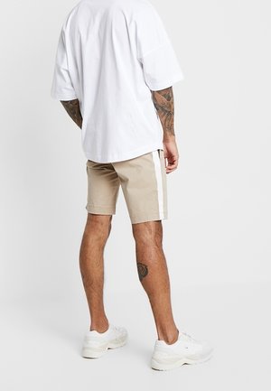 Uomo in piedi di spalle alla fotocamera, indossa una t-shirt bianca oversize, pantaloncini beige con banda laterale bianca, sneakers bianche e ha tatuaggi sulle braccia.