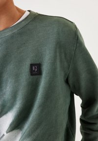 Felpa verde realizzata in tessuto morbido, con un collo a costine e una toppa del logo nero in rilievo sul fronte.