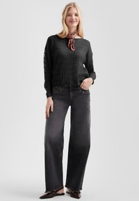 Vrouw in een zwart open-gehaakt sweater, donkere brede jeans met oogjesdetails, bruine puntige schoenen en een roze gepatterde sjaal om de nek.