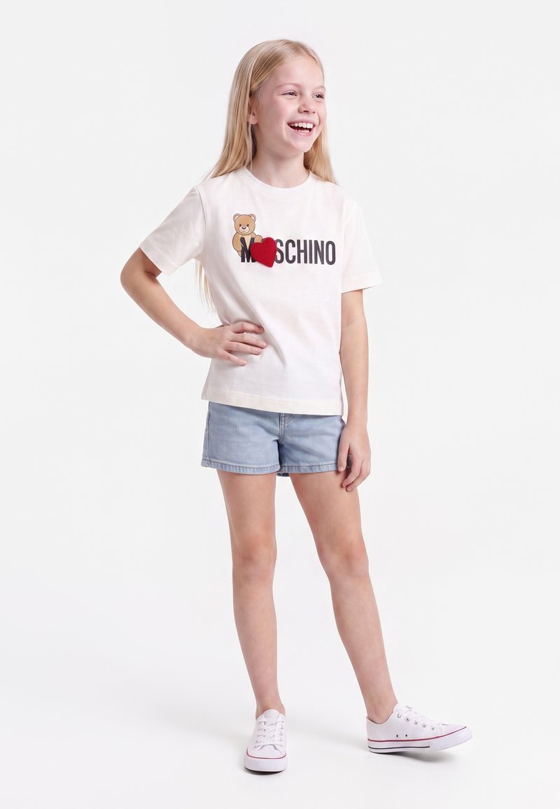 Млада момиче се усмихва с ръка на хълбока, облечена в бяла тениска Moschino с меченце и червено сърце, дънкови шорти и бели маратонки.