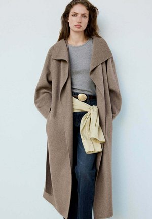 Mujer con abrigo largo marrón sobre camisa gris y vaqueros azules con un suéter beige atado alrededor de la cintura contra un fondo liso.