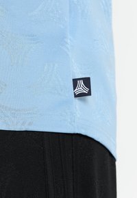 Ljusblått tyg med subtila geometriska mönster och en liten svart etikett med en vit triangulär logotyp, i kombination med en svart texturerad yta nedanför.