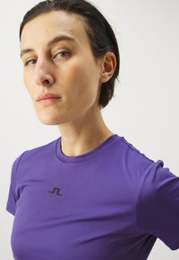 Violet sportinė marškinėlė su trumpomis rankovėmis, lygiais paviršiumi, su mažomis juodomis logotipo detalėmis krūtinėje ir peties srityje.