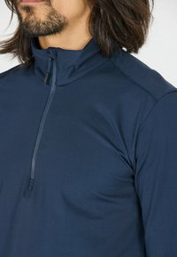 Marineblå atletisk pullover med høj krave og halv lynlås lukning. Har en glat overflade og diskret branding på skulderen.