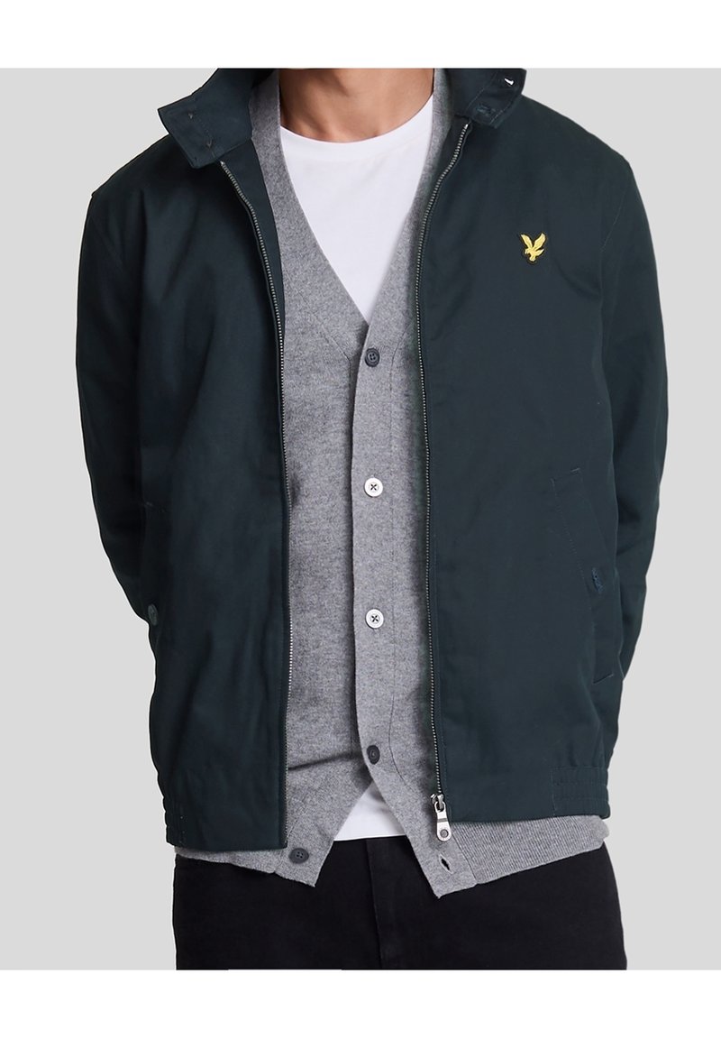 Navy zip-up jacket with a textured finish, featuring a pointed collar and a small embroidered logo. Worn over a gray cardigan and white shirt.