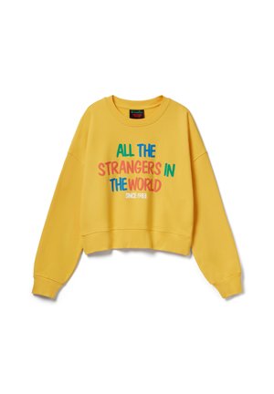Gele trui met lange mouwen en kleurrijke tekst "ALLE VREEMDELINGEN IN DE WERELD SINDS 1983" aan de voorkant, ronde hals en een ontspannen pasvorm.