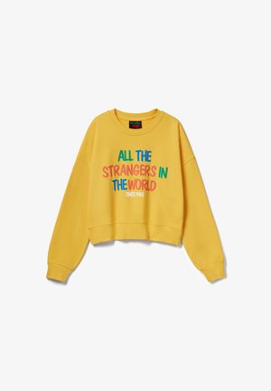 Gele trui met lange mouwen en kleurrijke tekst "ALLE VREEMDELINGEN IN DE WERELD SINDS 1983" aan de voorkant, ronde hals en een ontspannen pasvorm.