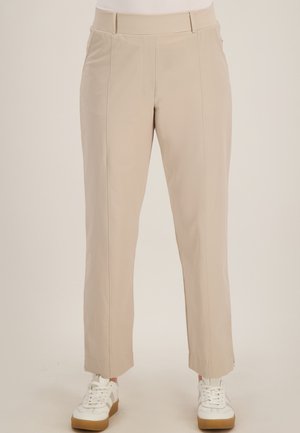 Beige op maat gemaakte broek met rechte pijpen en voorvouwen, gedragen met witte sneakers met rubberen zolen, getoond van taille tot voeten.