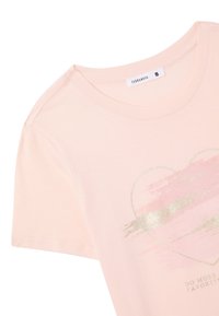 T-shirt rosa chiaro realizzato in morbido tessuto, con una grafica a forma di cuore con accenti dorati e design a pennellate, include il testo "FAI DI PIÙ I TUOI PREFERITI".