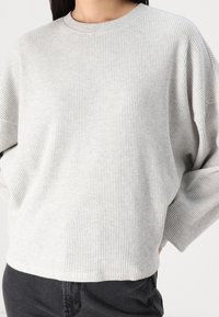 Maglione a coste grigio chiaro con scollo rotondo e spalle basse, abbinato a pantaloni neri. Tessuto morbido, vestibilità rilassata.